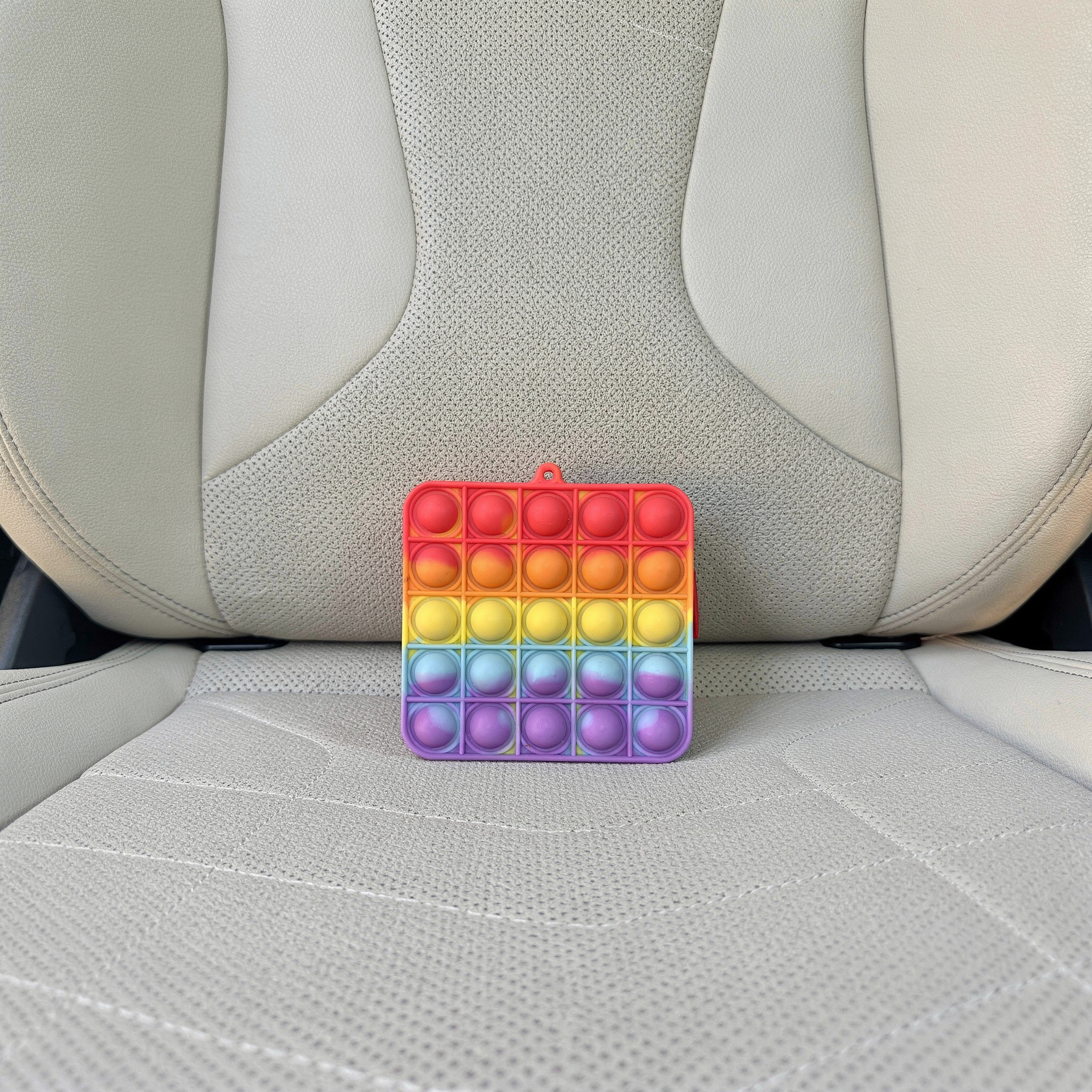 {
  "scene": "A lifestyle product shot placing the colorful square fidget toy from image 2 into the exact car seat setting from image 1. The environment is the interior of a car, featuring a light bei