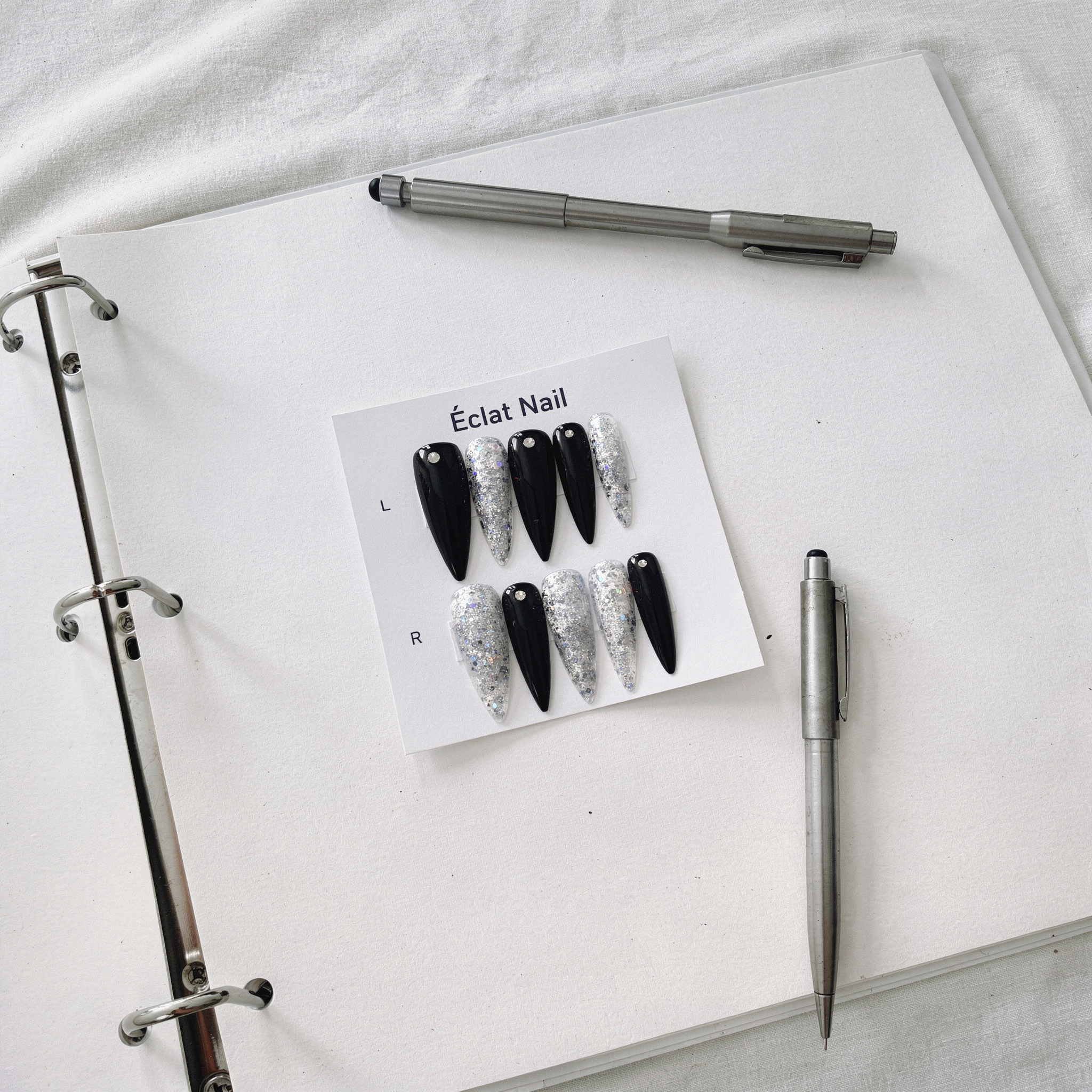 {
  "scene": "A top-down flat lay composition replacing the original item in image 1 with the false nail set from image 2. The scene features an open silver 3-ring binder with white paper resting on a