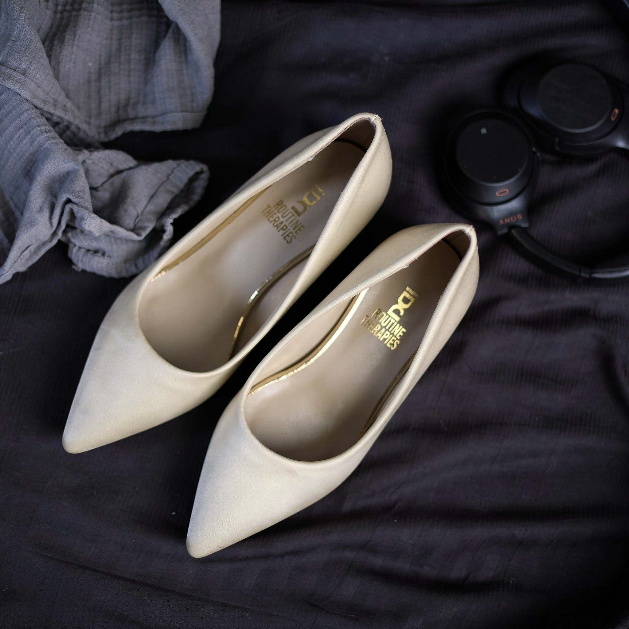 {
  "scene": "A top-down lifestyle flat lay photograph based on the setting of image 1, but with the central footwear replaced by the elegant pumps from image 2. The background is a dark, almost black