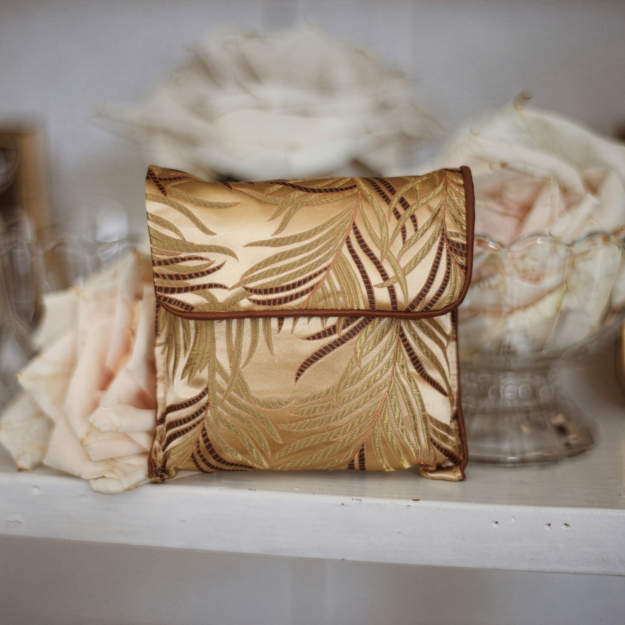 {
  "scene": "A product placement shot where the fabric pouch from image 2 is positioned centrally within the soft, elegant setting of image 1, replacing the original perfume bottle. The environment i
