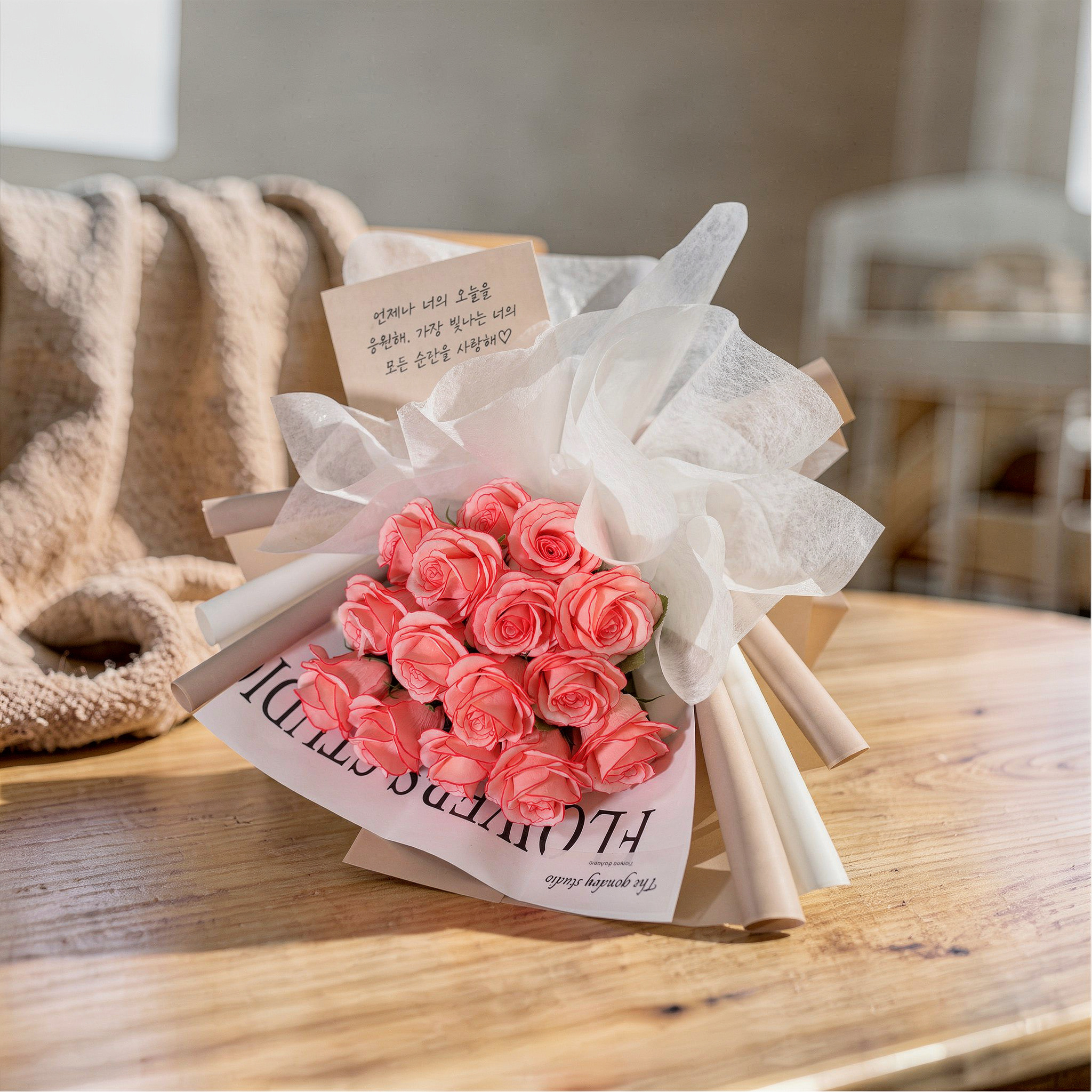 {
  "scene": "A product replacement shot where the metal cup in image 1 is replaced by the floral bouquet from image 2, placed centrally on the light-colored wooden table in a bright, airy indoor sett