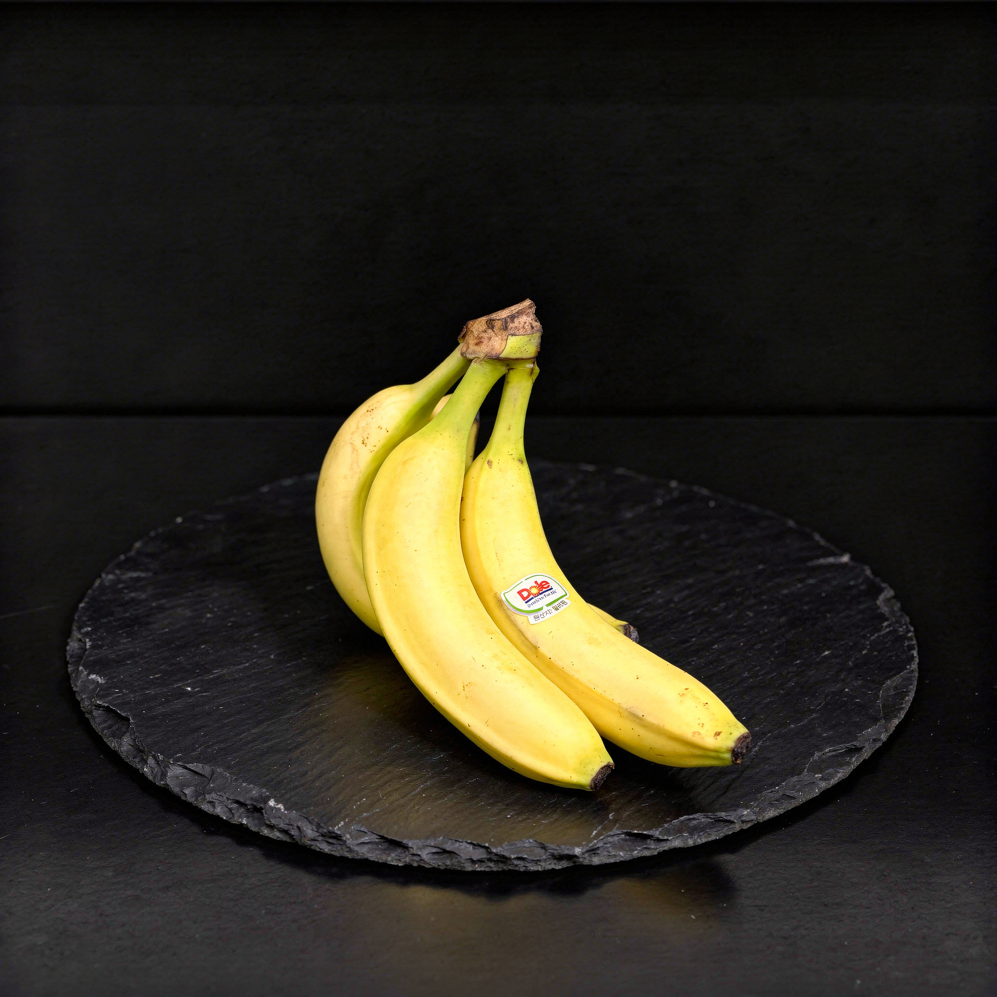 {
  "scene": "A premium commercial product photograph featuring the bunch of bananas from image 1 resting directly on a clean, dark slate kitchen countertop. The minimal, dark background provides high